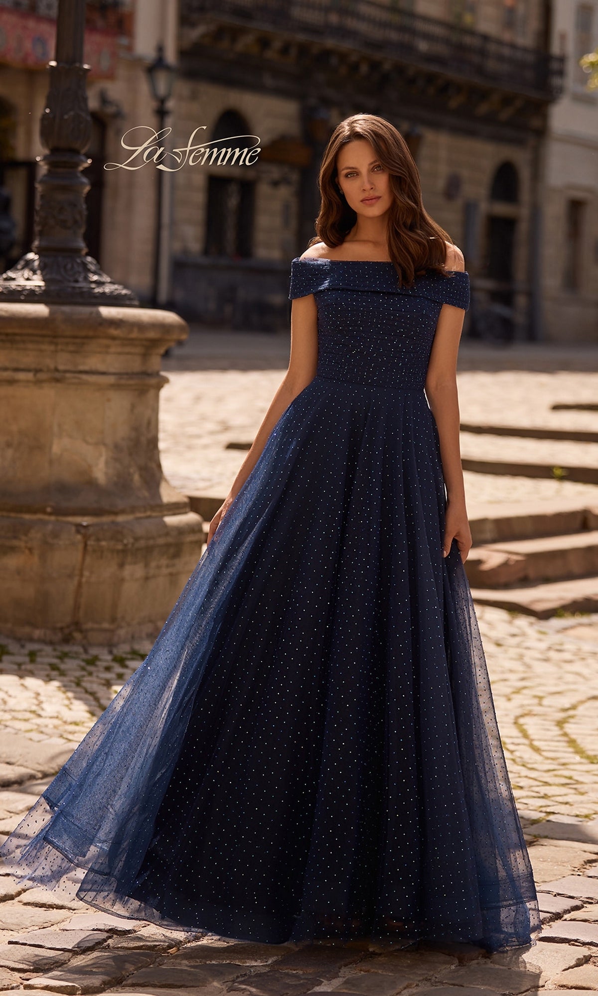 Woman in the Long Formal Dress: La Femme 28051, a navy off-the-shoulder gown with sparkles and full A-line skirt, stands on a cobblestone street by a stone lamp post.